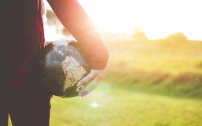 A photo taken from behind a woman standing while holding a globe on her hip looking out towards a sunny green field